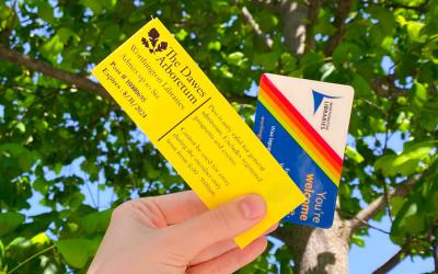 Hand holding Dawes Arboretum pass and library card against green leafy tree and blue sky